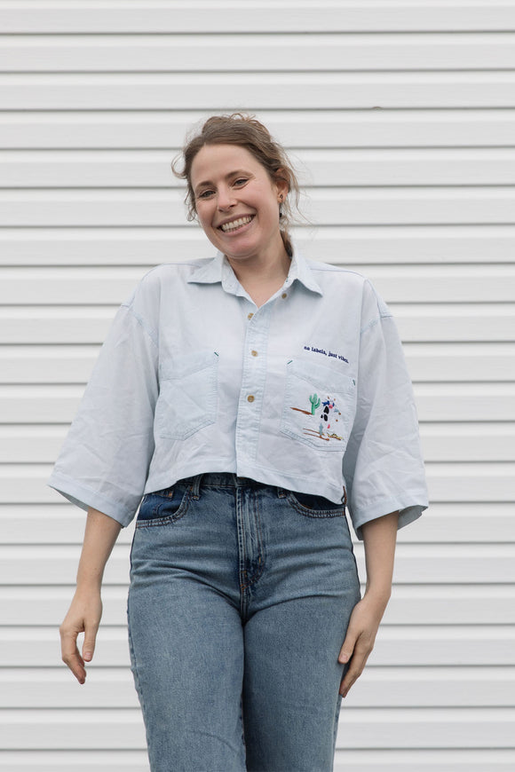 person wearing light blue cropped collared shirt with cow embroidery on breast pocket, upcycled in canada