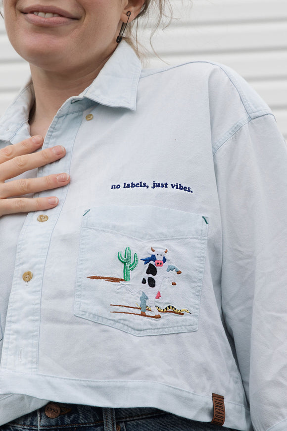 light blue chambray shirt, detail shot with cow, cactus and snake embroidery along with 'no labels, just vibes.'