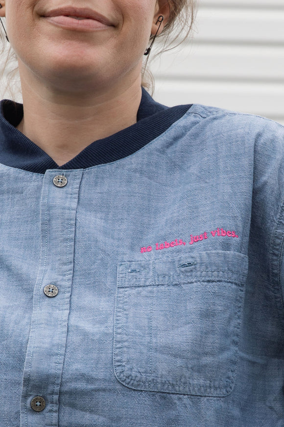 person wearing blue denim button up, upcycled in canada, detail shot featuring 'no labels, just vibes.' embroidery