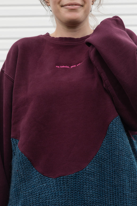 Person wearing a two-tone sweater with maroon and blue sections upcycled in canada, detail shot of 'no labels, just vibes.' embroidery