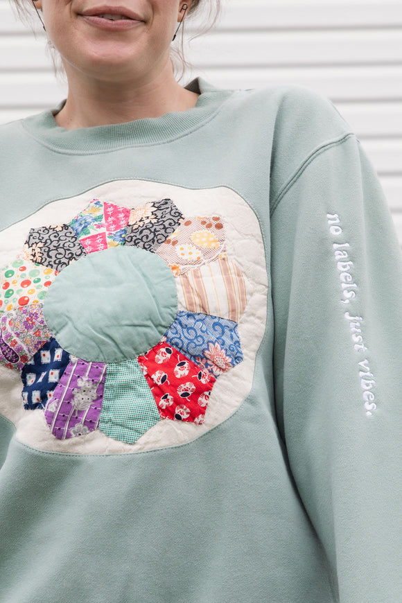 person wearing a mint green sweater with quilted pinwheel on the front, upcycled in canada, detail shot of 'no labels, just vibes.' embroidery