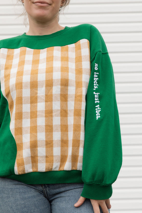 Person wearing a green and plaid sweater, upcycled in canada, detail shot of 'no labels, just vibes.' embroidery