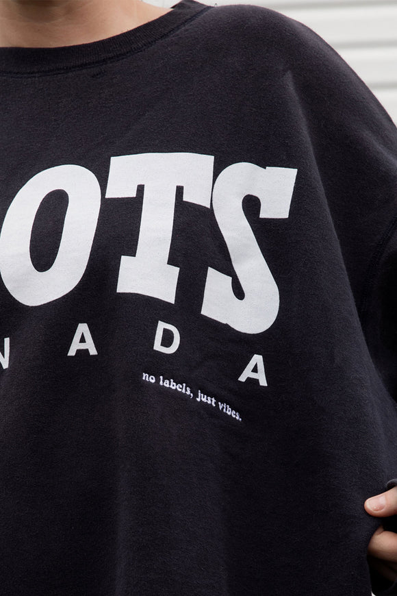 Person wearing a black 'ROOTS Canada' sweatshirt, upcycled in canada, detail shot of 'no label, just vibes.' embroidery