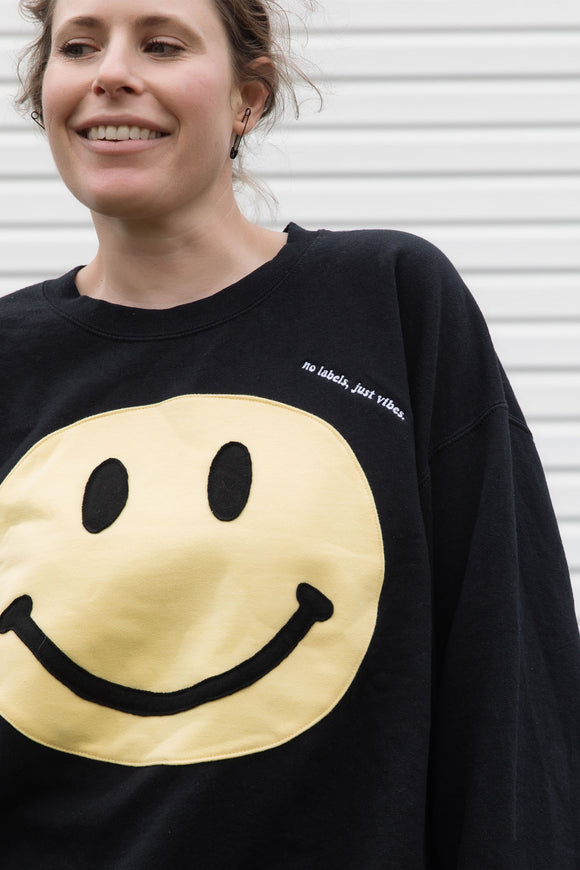 Person wearing a black sweatshirt with a yellow smiley face design, upcycled in canada, detail shot of 'no labels, just vibes.' embroidery