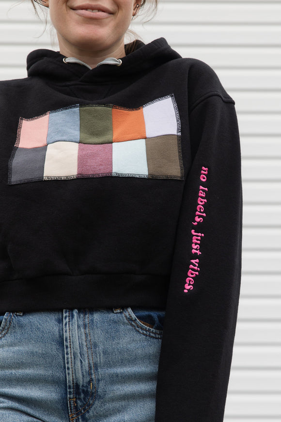 Person wearing a black hoodie with a colorful square design, upcycled in canada, featuring 'no labels, just vibes.' embroidery on the sleeve