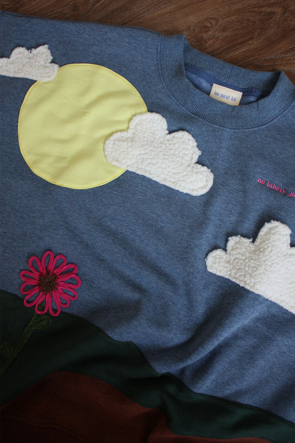 upcycled earth sweater featuring the earth, grass, sky with the sun, clouds and a flower, repurposed in canada, detail shot of 'no labels, just vibes.' embroidery