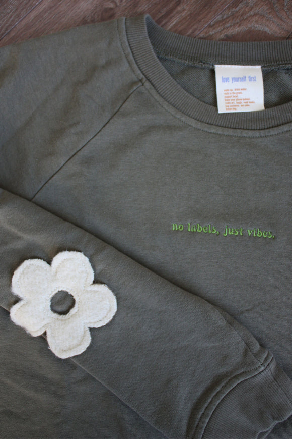 green sweater with cream flowers down the sleeves, upcycled in canada, detail shot of 'no labels, just vibes.' embroidery