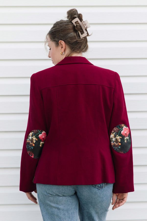 A woman viewed from the back wearing a deep burgundy blazer with floral embroidered elbow patches and light blue jeans, standing against a white horizontal siding wall.