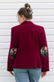 A woman viewed from the back wearing a deep burgundy blazer with floral embroidered elbow patches and light blue jeans, standing against a white horizontal siding wall.