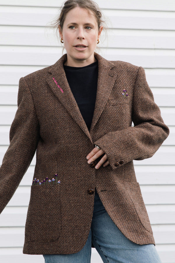 A woman wearing a brown textured tweed blazer with colorful embroidered details on the lapels and pockets, paired with a black top and blue jeans, standing against a white siding background.