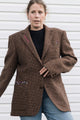 A woman wearing a brown textured tweed blazer with colorful embroidered details on the lapels and pockets, paired with a black top and blue jeans, standing against a white siding background.