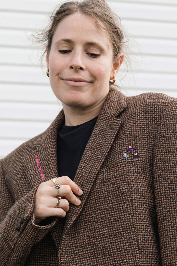 A woman wearing a brown tweed blazer over a black top, featuring a small purple and pink floral brooch on the lapel, set against a white paneled background.