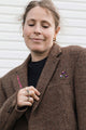 A woman wearing a brown tweed blazer over a black top, featuring a small purple and pink floral brooch on the lapel, set against a white paneled background.