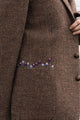 Close-up of a brown textured tweed coat with a decorative embroidered row of small purple, blue, and pink flowers across the pocket, shown on a model.
