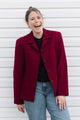 A woman laughing outdoors in front of a white siding wall, wearing a dark red tailored blazer over a black top and light blue jeans.