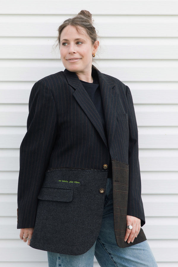 A woman wearing a black pinstriped blazer with unique patch pockets over a black top and blue jeans, standing against a white horizontal siding wall.