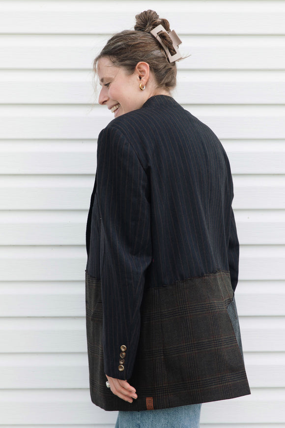 A woman wearing a dark navy pinstriped blazer with brown and black plaid patch pockets, photographed from the back against a white horizontal siding wall.