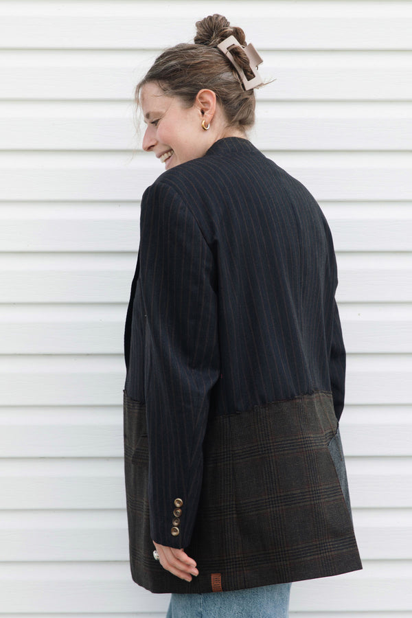 A woman wearing a dark navy pinstriped blazer with brown and black plaid patch pockets, photographed from the back against a white horizontal siding wall.