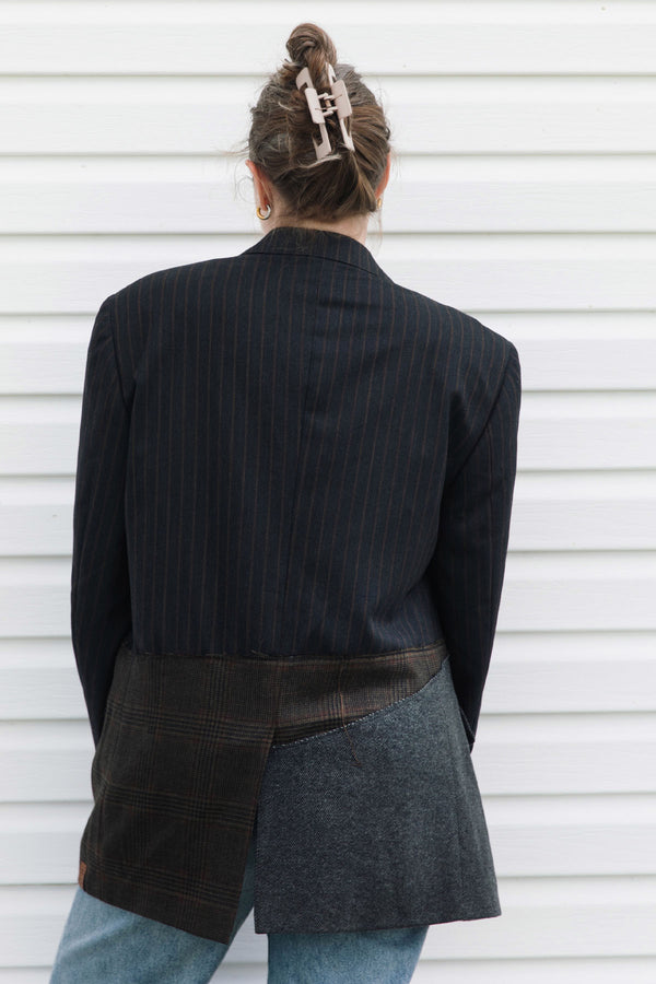 A person shown from the back wearing a dark navy pinstripe blazer over a patchwork skirt with brown plaid and gray wool-like panels, paired with blue jeans, standing against a white horizontal siding wall.