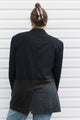 A person shown from the back wearing a dark navy pinstripe blazer over a patchwork skirt with brown plaid and gray wool-like panels, paired with blue jeans, standing against a white horizontal siding wall.