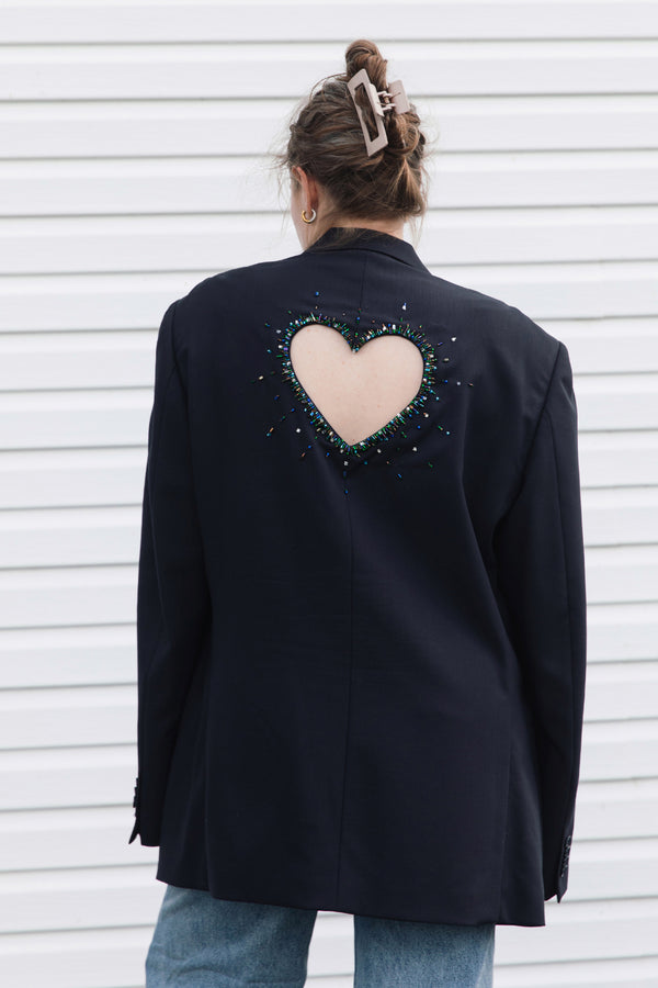 A woman viewed from behind wearing a dark navy blazer with a large heart-shaped cutout on the back, embellished with colorful rhinestones, paired with light blue jeans against a white horizontal siding background.