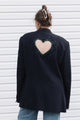 A woman viewed from behind wearing a dark navy blazer with a large heart-shaped cutout on the back, embellished with colorful rhinestones, paired with light blue jeans against a white horizontal siding background.