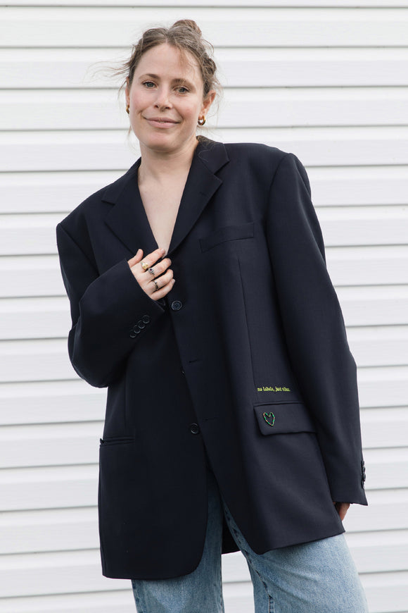 A woman wearing an oversized black blazer with light blue jeans, standing against a white horizontal-slat wall. The blazer features a subtle green embroidered logo on the pocket flap.