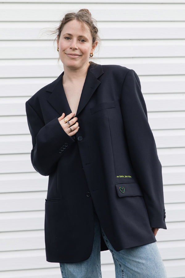 A woman wearing an oversized black blazer with light blue jeans, standing against a white horizontal-slat wall. The blazer features a subtle green embroidered logo on the pocket flap.