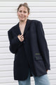 A woman wearing an oversized black blazer with light blue jeans, standing against a white horizontal-slat wall. The blazer features a subtle green embroidered logo on the pocket flap.