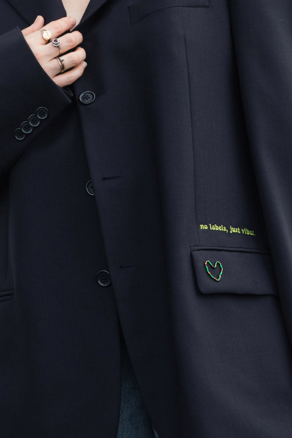 Close-up of a dark navy blazer with a pocket featuring a green embroidered heart and yellow text reading 'no labels, just style.' A hand with rings is visible near the collar.