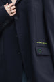 Close-up of a dark navy blazer with a pocket featuring a green embroidered heart and yellow text reading 'no labels, just style.' A hand with rings is visible near the collar.