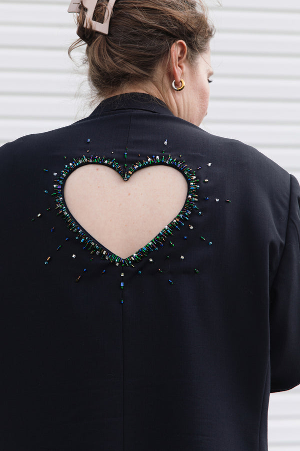 A woman shown from behind wearing a black jacket with a large heart-shaped cutout on the back, embellished with a border of multicolored rhinestones and beads.
