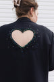 A woman shown from behind wearing a black jacket with a large heart-shaped cutout on the back, embellished with a border of multicolored rhinestones and beads.