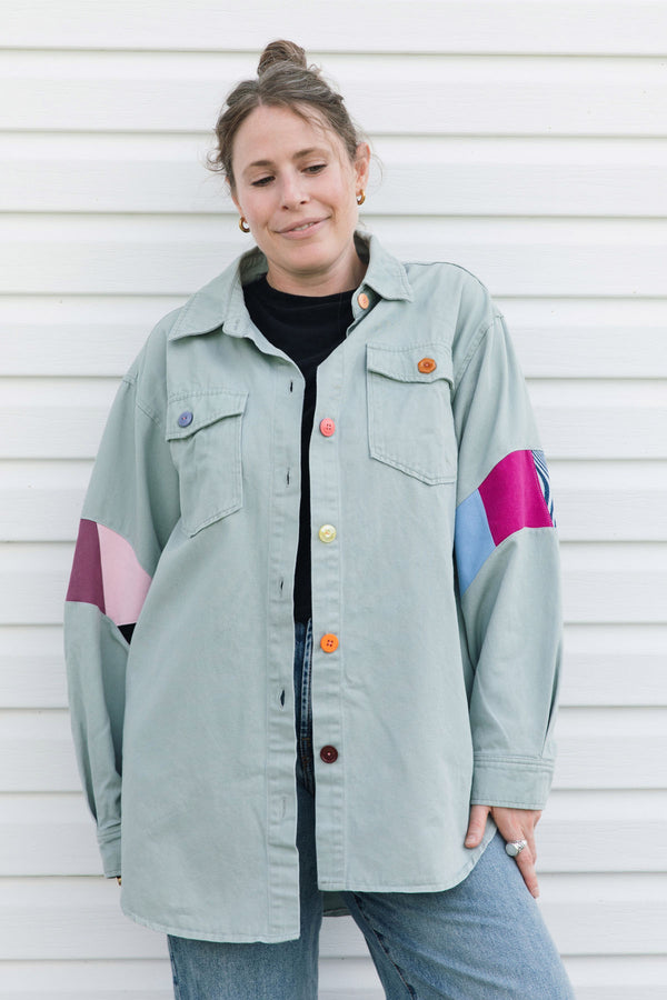 A woman wearing a sage green button-up shirt jacket with colorful geometric color-blocked panels on the sleeves, styled with blue jeans and a black t-shirt, standing against a white siding wall.