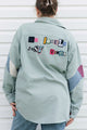 A person viewed from behind wearing a sage green button-up shirt with colorful patchwork lettering on the back reading 'DO LABEL JUST VIOLES' and patterned sleeve panels, paired with blue jeans against a white siding background.