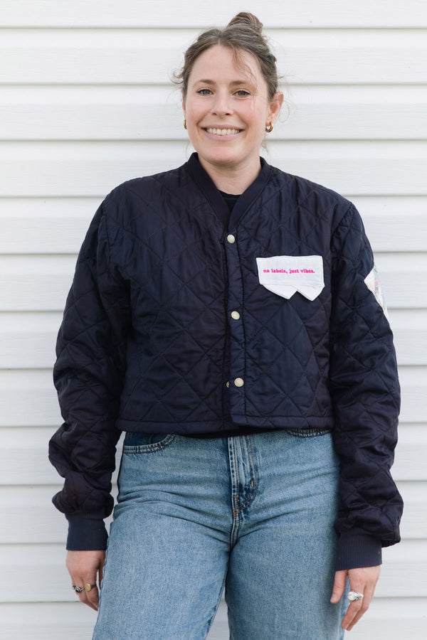 A woman wearing a navy blue quilted bomber jacket with a white chest patch that reads 'no bullshit, just clothes' and light blue jeans, standing against a white horizontal siding background.