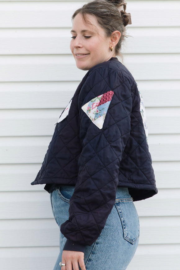 A woman wearing a navy blue quilted bomber jacket with colorful patchwork details on the chest and sleeves, paired with blue jeans, standing against a white paneled wall.