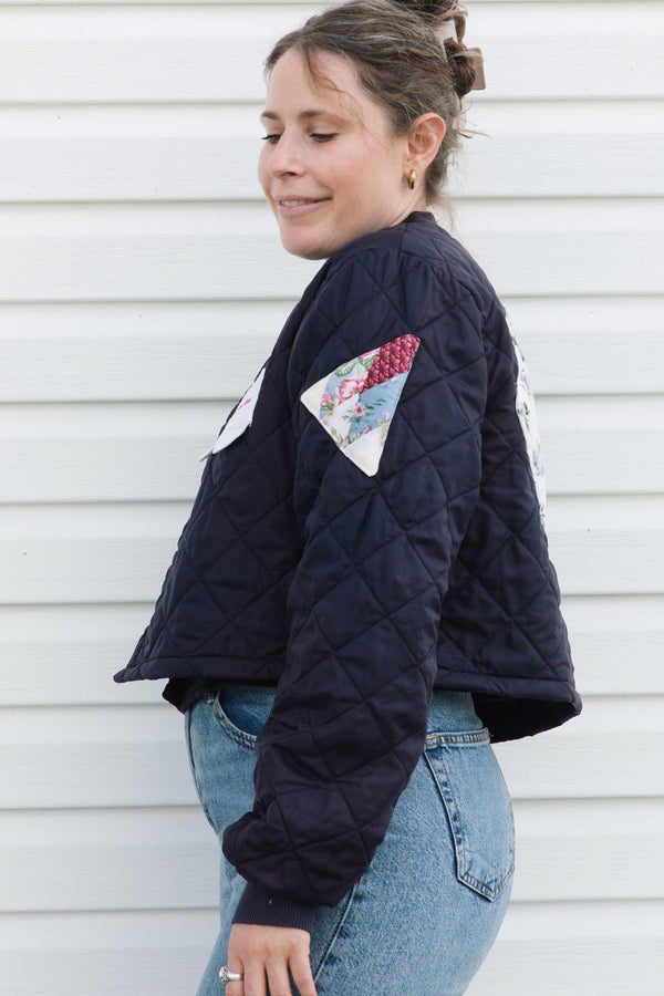 A woman wearing a navy blue quilted bomber jacket with colorful patchwork details on the chest and sleeves, paired with blue jeans, standing against a white paneled wall.