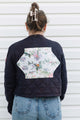 A woman viewed from behind wearing a navy quilted bomber jacket with a large hexagonal floral patch on the back, paired with blue jeans against a white siding background.