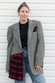 A woman stands against a white siding wall wearing an oversized gray plaid blazer over a black t-shirt and blue jeans, with a red and black plaid pocket square and matching plaid draped over one leg.