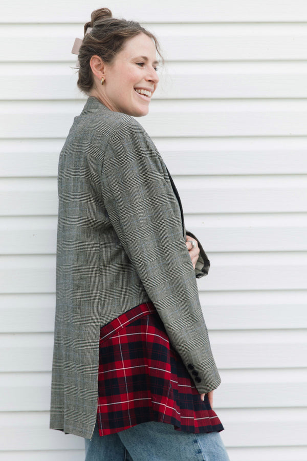 A woman wearing an oversized gray plaid blazer over a red and black plaid shirt, paired with blue jeans, standing against a white siding wall.
