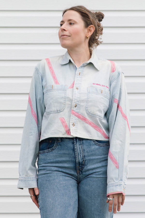 A woman wearing a light-wash denim button-up shirt with pink diagonal stripe patterns and blue jeans, standing against a white horizontal-slat wall.