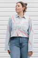 A woman wearing a light-wash denim button-up shirt with pink diagonal stripe patterns and blue jeans, standing against a white horizontal-slat wall.