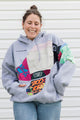 A woman wearing a light gray oversized hoodie with a colorful graphic patchwork design featuring a large Mickey Mouse silhouette and various vibrant text and pattern panels, paired with blue jeans, standing in front of a white siding wall.