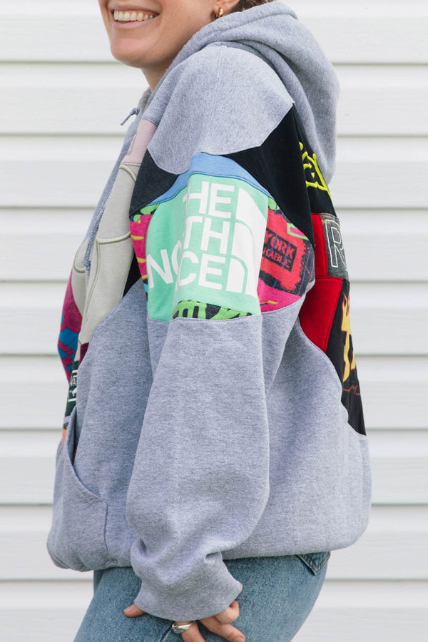 A person wearing a light gray hoodie with large multicolor graphic patch panels on the sleeves, paired with blue jeans, standing against a white siding background.