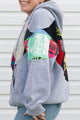 A person wearing a light gray hoodie with large multicolor graphic patch panels on the sleeves, paired with blue jeans, standing against a white siding background.
