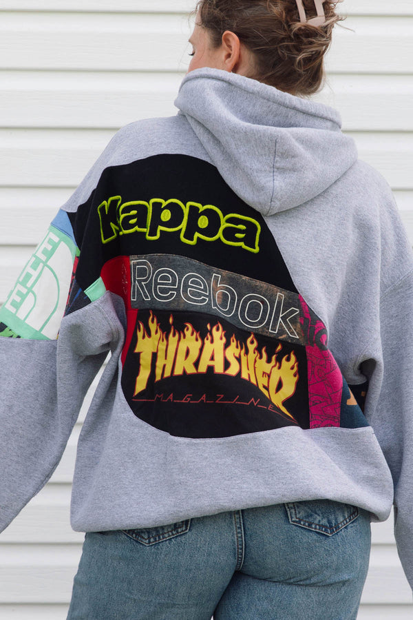 A person wearing a light gray hoodie from the back, featuring a large graphic patch with 'Kappa', 'Reebok', and 'Thrasher' logos. The person is also wearing blue jeans, standing against a white paneled wall.