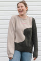 A woman smiling and wearing a color-block knit sweater with a beige body, dark gray textured sleeves and lower panel, and light blue jeans, standing against a white siding background.