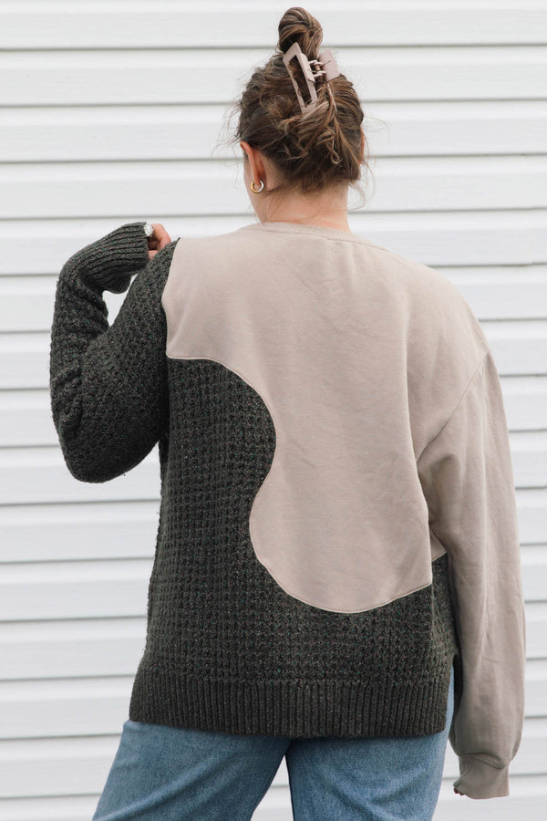 A person shown from the back wearing a color-block knit sweater with a dark charcoal gray textured body and sleeves, and a large beige back panel, paired with blue jeans against a white siding background.