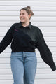 A woman smiling and posing in a black asymmetrical long-sleeve button-up shirt with light blue jeans, standing against a white horizontal siding background.